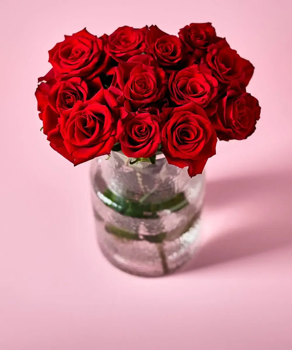 A bright bouquet of yellow roses, white blooms, and greenery arranged in a ribbed white vase, with additional flowers displayed in an open box behind it on a soft pink background.